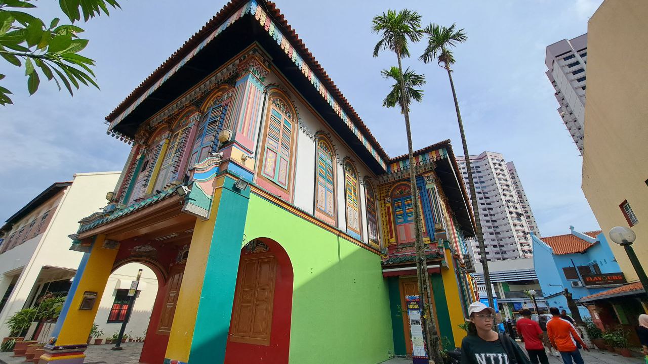 a vibrant and colorful shophouse stands out on a busy street in singapore. the building is adorned with intricate details and painted in a rainbow of hues, creating a striking contrast against the surrounding buildings. the shophouse's facade is a testament to the rich architectural heritage of singapore, showcasing a blend of traditional and modern elements.  a glimpse of other buildings can be seen in the background, including modern highrises and a traditional blue shophouse.  the scene captures the lively and eclectic atmosphere of singapore, a city that blends historical charm with modern dynamism.