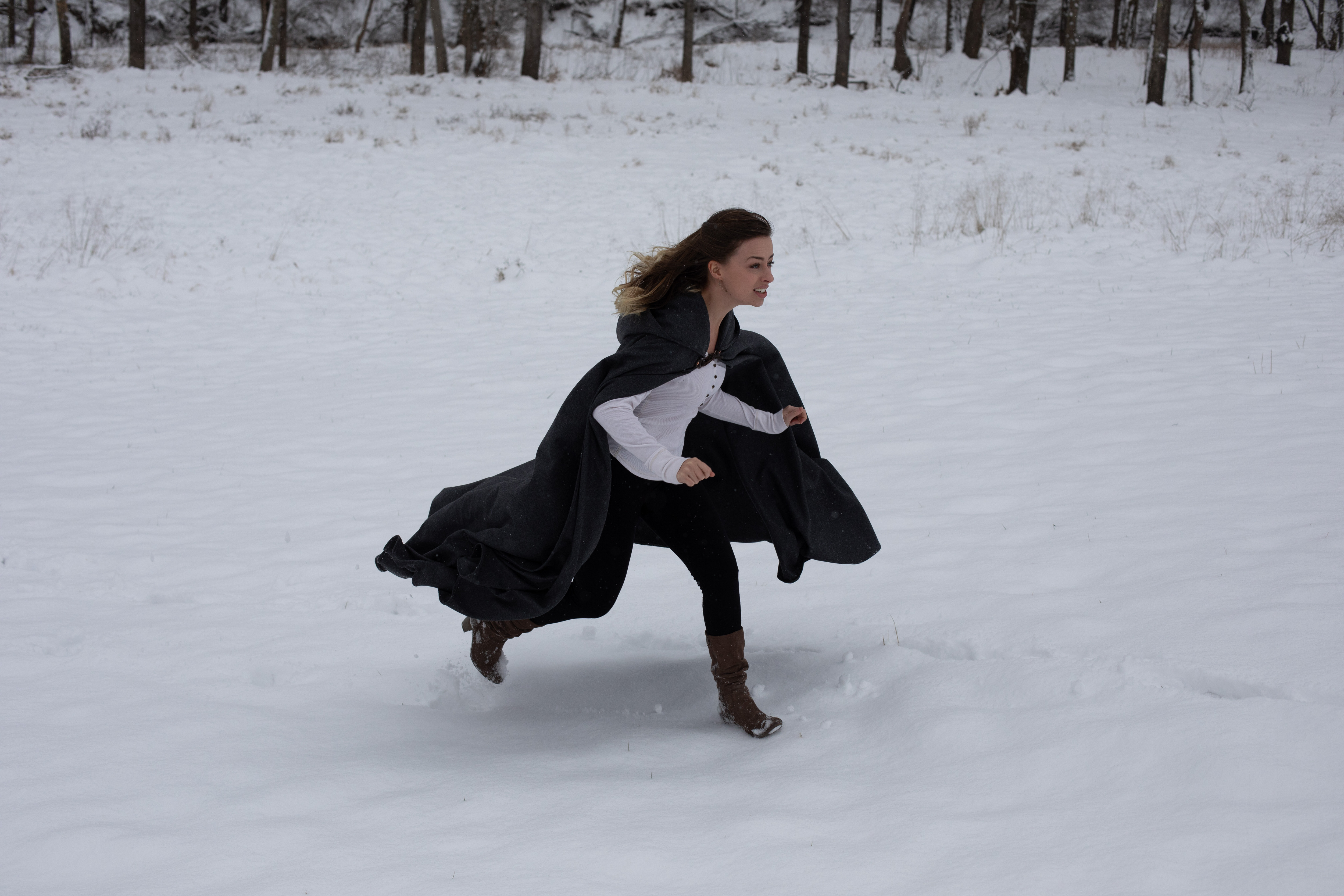 a young woman in a black cape runs through the snow in a field. the image evokes a sense of freedom, adventure, and fantasy.  the background is a snowy forest with bare trees.  the overcast sky provides soft, natural lighting.