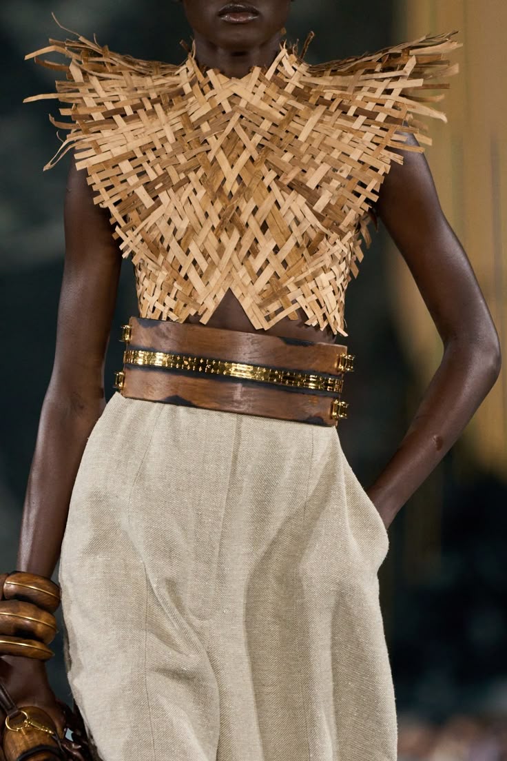 a darkskinned model is shown on a runway, likely during a fashion show. she is wearing a woven strawlike top with a triangle cutout at the center of the bust. the top has a high collar and extends slightly over the shoulders. the model is also wearing highwaisted, beige, linenlike pants and a wide brown belt with gold embellishments around the waist. on one wrist, she has a set of thick, rounded wooden bracelets. her hand is placed in her pocket. the overall aesthetic is earthy and natural.