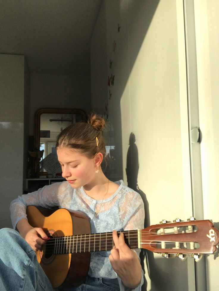 a young woman sits in a room, playing an acoustic guitar. she is wearing a blue shirt and jeans. the sunlight is coming through the window behind her, illuminating her hair and the guitar. she is looking down at the guitar as she plays.