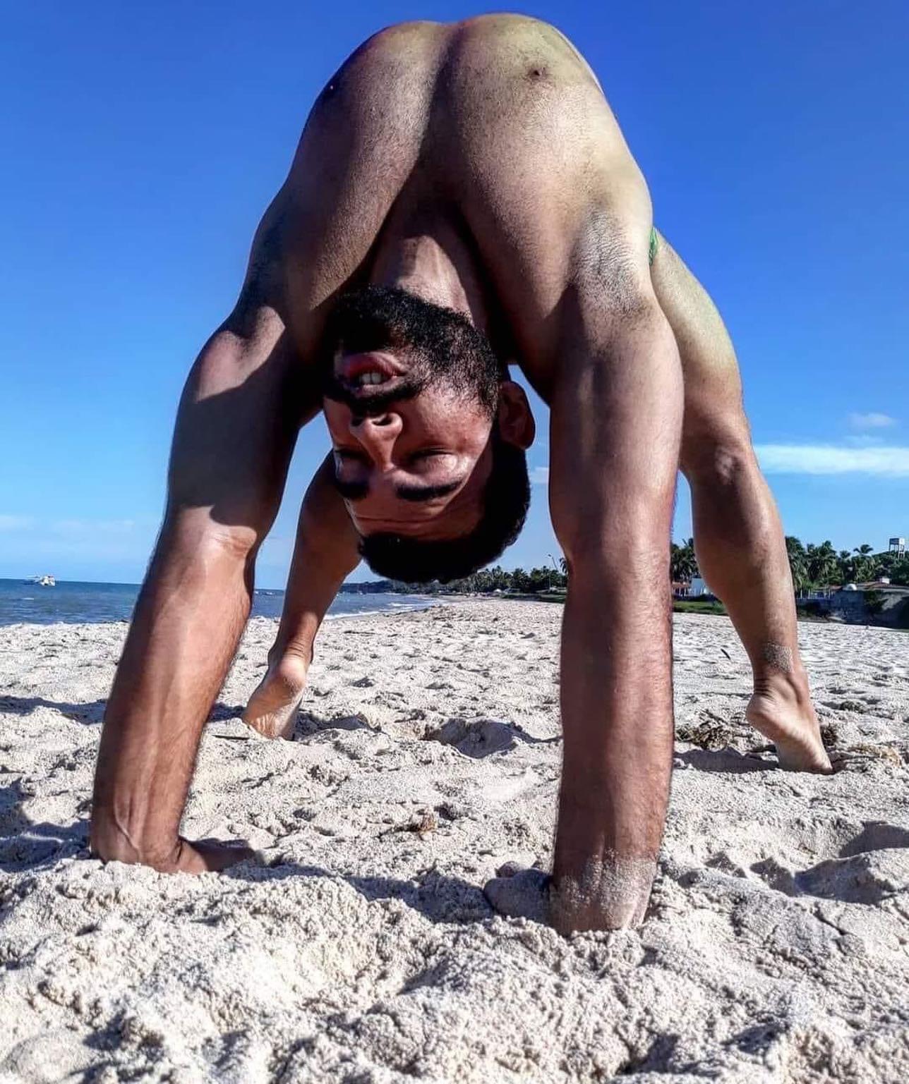 a muscular man with dark hair and beard is performing an impressive contortionist yoga pose on a sunny beach. his body forms an arch, with his hands and feet planted firmly on the sand, his head upside down, and his gaze directed towards the camera. the blue sky, white sand, and glistening ocean create a vibrant backdrop for this display of strength and flexibility.