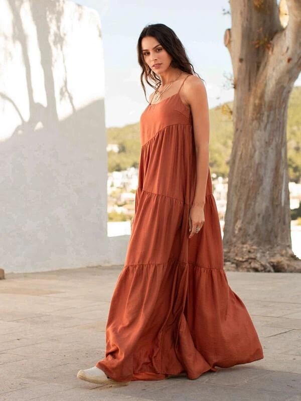 a woman is standing outdoors in a long terracottacolored tiered maxi dress with spaghetti straps and wearing espadrilles. she is wearing a necklace. she is standing near a large tree and a white wall, with a mediterraneanstyle building in the background.