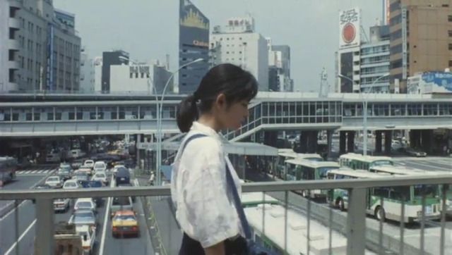 a young asian woman stands on a pedestrian bridge overlooking a busy street in a japanese city.  numerous cars and buses fill the road below.  tall buildings and skyscrapers form the background, creating a vibrant cityscape. the image has a retro feel, possibly from the 1980s or 1990s. the woman is the focal point, wearing a lightcolored shirt and looking towards the street below. the overall impression is one of bustling urban life in japan.