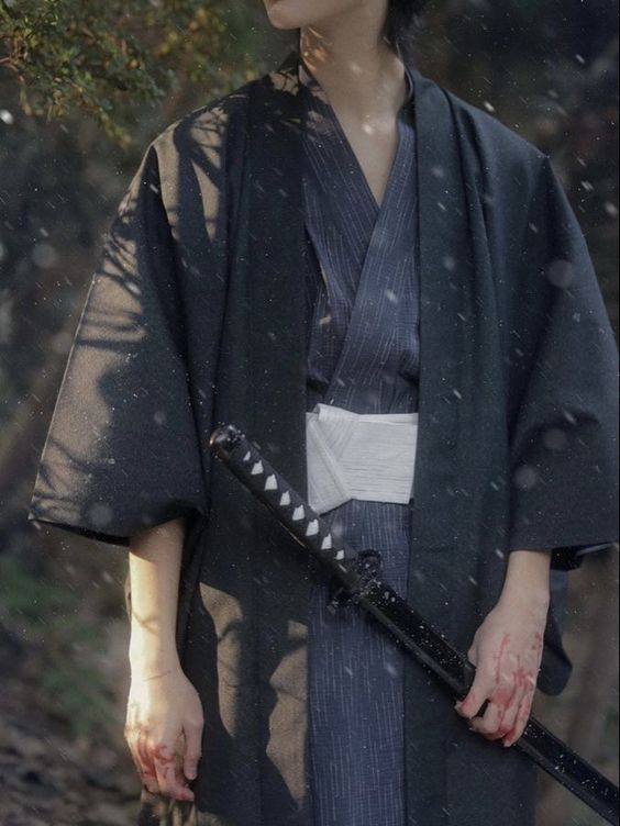 a young woman dressed as a samurai with a sword.  the image is a close up of her torso as she stands in a forest with dappled sunlight. her hands are stained with blood.