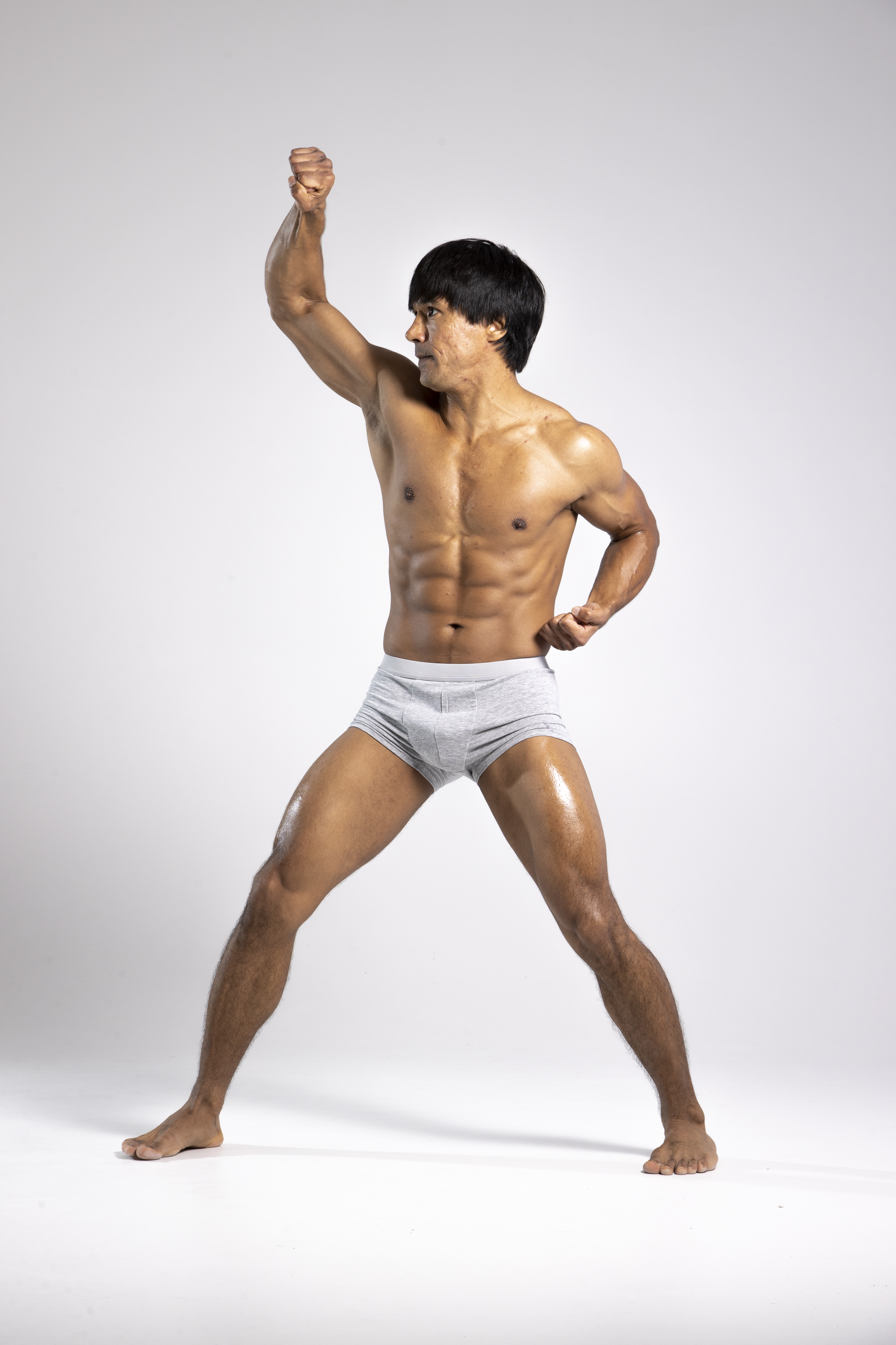 a muscular, shirtless asian man in gray underwear poses in a martial arts stance against a white studio background. his right arm is raised with a clenched fist, and his left hand is gesturing near his waist. he is barefoot, and his physique is welldefined.