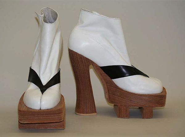 two white platform boots are standing sidebyside on a light surface. the left boot is viewed from the front and shows a black vshaped strap. the right boot is viewed from the side and shows a solid black strap. both boots have a wood platform and heel.