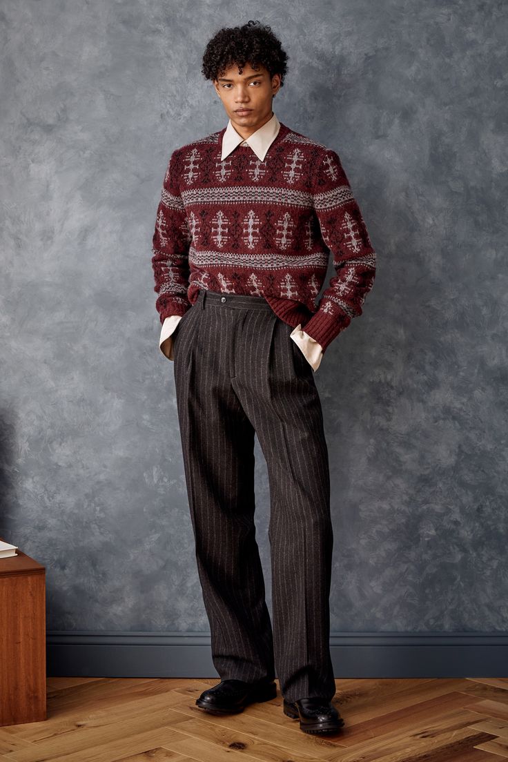 a fullbody shot of a male model standing against a dark gray textured wall. he's wearing a burgundy fair isle patterned sweater over a white collared shirt, dark gray pinstripe pleated trousers, and black oxfords. his hands are in his pockets. the floor is a light brown wood.  the background is a simple studio setup. the overall style is smart casual.