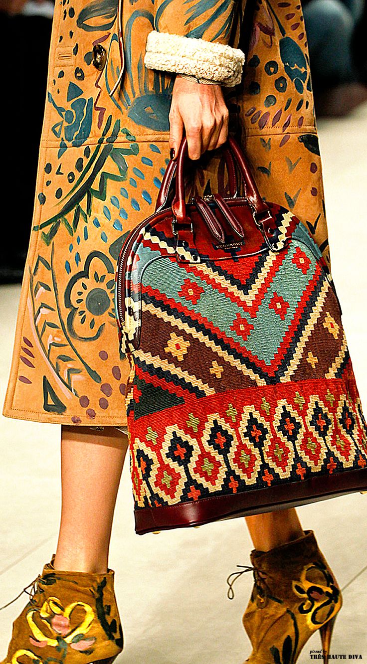 closeup shot of a woman's lower body showcasing a stylish autumn/winter outfit. she's wearing a long, suede coat with handpainted, colorful floral and abstract patterns.  the coat has a sheepskin or fur trim on the cuffs. her handbag is a striking piece; a rich burgundy satchel with a woven, intricately patterned front that seems to have ethnic or bohemian inspiration. the vibrant colors in the bag complement the coat’s design. she's also wearing brown ankle boots with painted details similar in style to the coat. the overall aesthetic is bohemianchic with highend luxury elements. the image is likely a fashion runway shot.