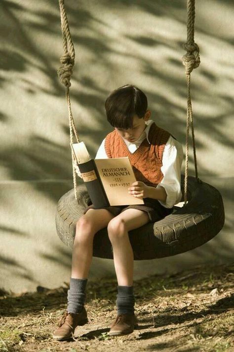 a young boy wearing a white shirt and orange sweater is sitting on a tire swing holding an old book and reading. he is very focused on his reading. he is wearing dark brown shoes and dark socks. he is sitting outside on a patch of green grass and dirt. he is very engrossed in the book he is holding. there are shadows in the background on a wall. the book in his hand is german and seems to be an almanac.