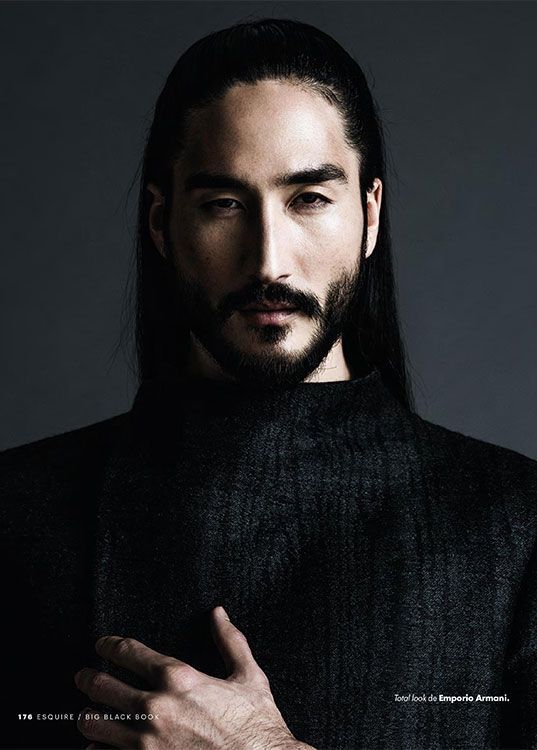 a portrait of a male model with long black hair, a beard, and a mustache. he is wearing black clothing and standing against a dark background. he has a serious expression and his hand is on his chest. the image is from esquire's big black book and features a total look by emporio armani.