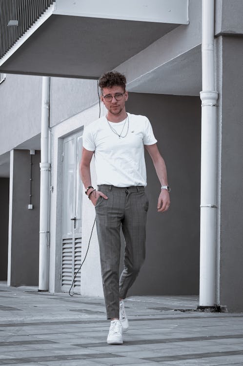a young, stylish man walks down the street. he is wearing a white tshirt, grey trousers, and white sneakers. his outfit is simple yet chic, and he exudes a confident and relaxed vibe. the urban setting provides a neutral backdrop that complements his stylish clothing choices. the image overall conveys a sense of effortless cool.