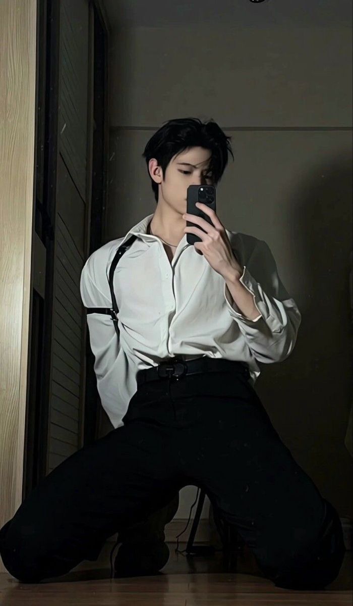 a young man stands in a dimly lit room, posing for a selfie. he's wearing a white buttondown shirt, black pants, and black shoes. he's holding his phone up to his face and staring directly at the camera.  he appears serious and stylish.