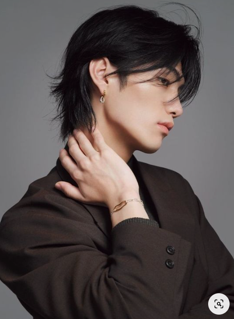 the image portrays a stylish young man in a closeup, side profile shot. he has mediumlength, dark hair styled with a side part and a slight wave. he is wearing a dark brown suit jacket and silver jewelry  a small hoop earring and a delicate bracelet with a pendant. the background is a plain, neutral gray. the lighting is soft and flattering, highlighting his features and the texture of his hair.  the image evokes a sense of modern fashion and sophisticated style.