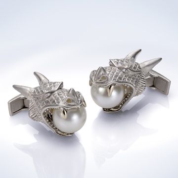 the image shows a pair of silver cufflinks in the shape of dragon heads, each with a pearl held in its mouth. the cufflinks are on a reflective surface.