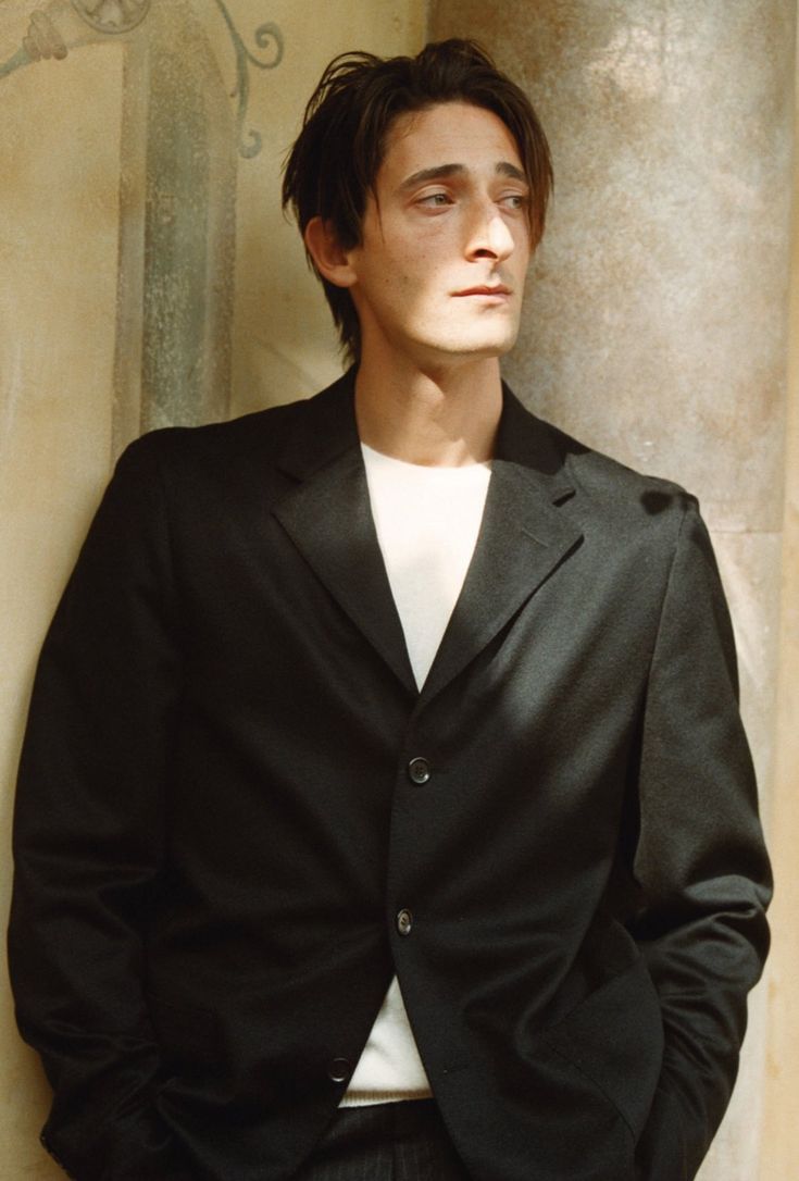 the image is a portrait of a man. he is standing against a textured wall or column that looks like it may be from an old building or have a stuccolike appearance. the man is wearing a darkcolored, possibly black, blazer over a white tshirt. his hair is dark brown and slightly tousled. his expression is serious and somewhat pensive. he is looking off to the side and the light casts shadows on his face and jacket. the overall tone is sophisticated and elegant, possibly suggesting a fashion photograph.