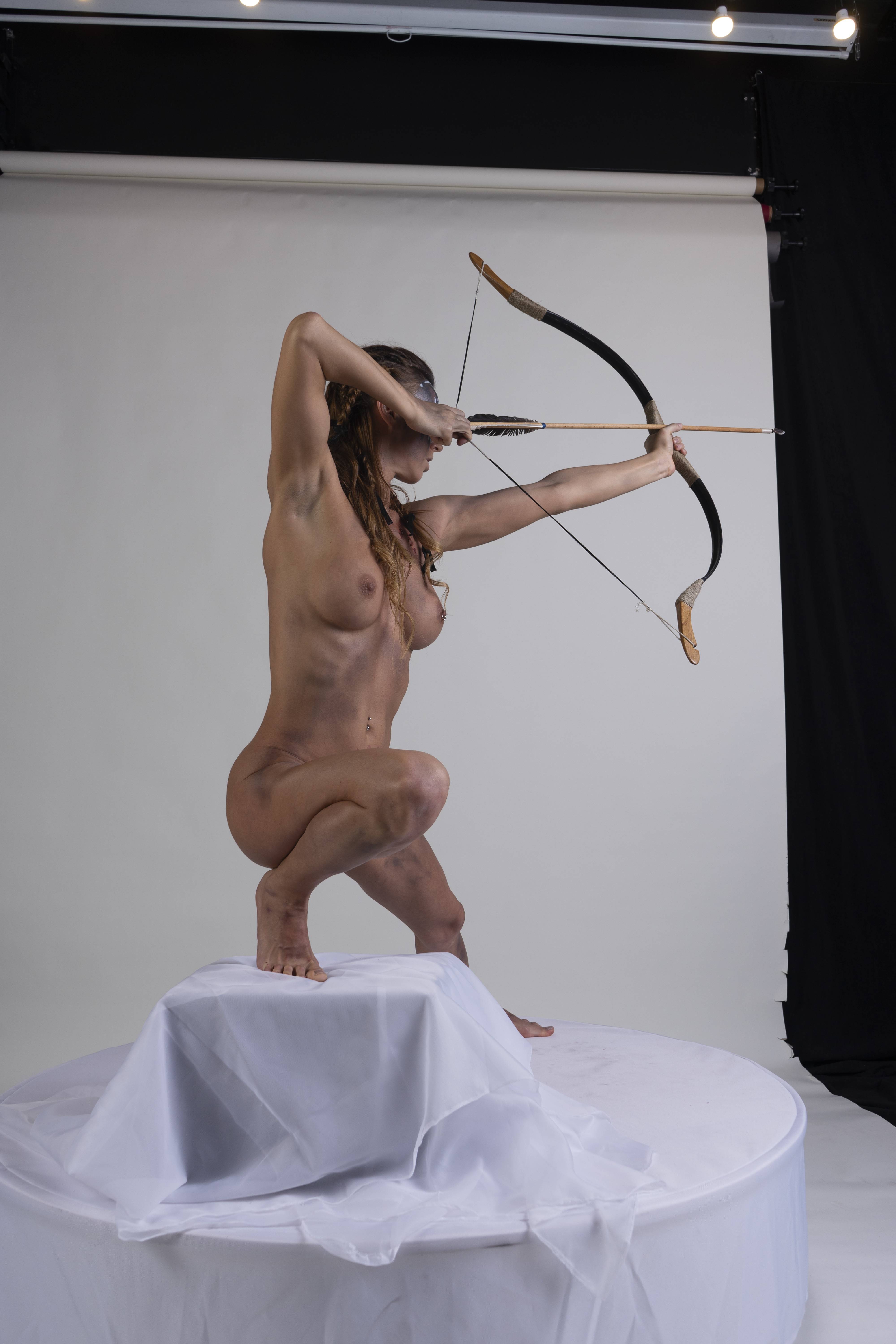 a nude, fairskinned woman with braids stands crouched on a white clothcovered platform, aiming a bow and arrow. she is in a studio setting, and her body is adorned with what appears to be mud or body paint in a tribal style. her muscular build and focused expression suggest she is a warrior or hunter. the lighting equipment is visible in the background.