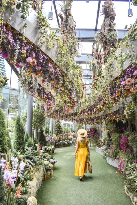a woman in a yellow dress and straw hat walks down a path in a lush indoor garden.  the path is lined with vibrant flowers and plants, and overhead, hanging floral arrangements create a stunning canopy. the architecture of the building is modern and incorporates large glass panels. the scene is bright, colorful, and peaceful.