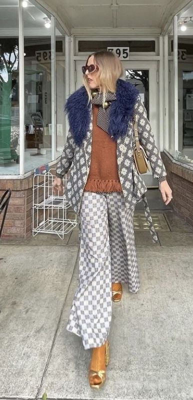a woman in a stylish retro outfit stands outside a brick building. her outfit features a checkered pair of widelegged pants, a floral jacket with a dark blue fur collar, a burnt orange sweater, and gold sandals. she is carrying a brown handbag and wearing sunglasses. the overall style evokes a 70s or 60s vibe.