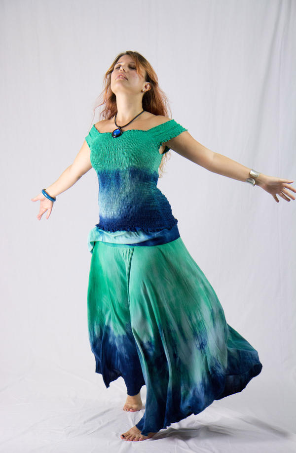 a full shot of a woman with long, wavy reddishblonde hair wearing a flowing teal and blue tiedye dress. the dress has a smocked bodice and a long, full skirt. she is barefoot and her arms are outstretched, conveying a sense of peace and freedom.  she wears simple jewelry – a necklace and bracelets – and her eyes are closed, suggesting a meditative or spiritual state.  the background is plain white, focusing attention on the model and her outfit. the overall style is bohemian or hippieinspired.