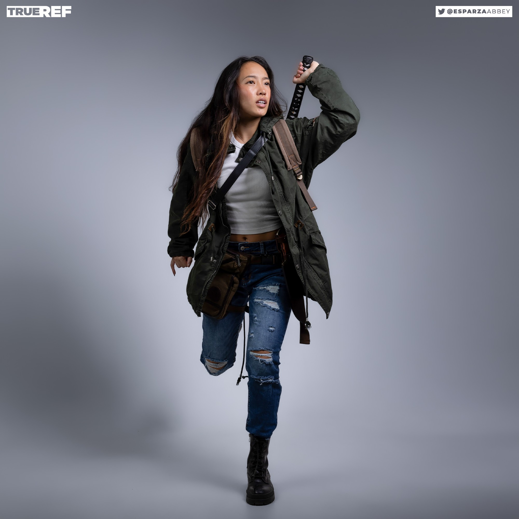 a young woman, possibly cosplaying or in a character portrayal, is captured in a dynamic running pose against a neutral grey backdrop. she's dressed in a dark green parka, ripped jeans, and combat boots, carrying a katana over her shoulder and a backpack. her determined expression and energetic posture suggest a narrative of action and resilience. the image is professionally shot, showcasing a polished aesthetic.