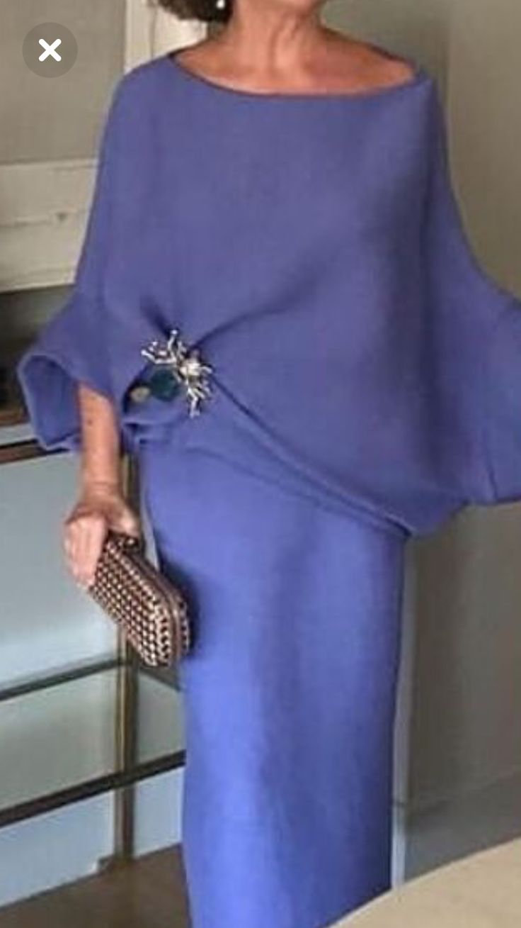 the image shows a woman wearing a stylish, loosefitting, periwinkle or light purple midi dress with wide sleeves and a bateau neckline. the dress has a relaxed, almost draped silhouette. she is accessorized with a statement brooch on her chest and a textured clutch. the setting appears to be indoors, possibly in a welllit home or boutique.  the overall impression is one of sophisticated elegance.