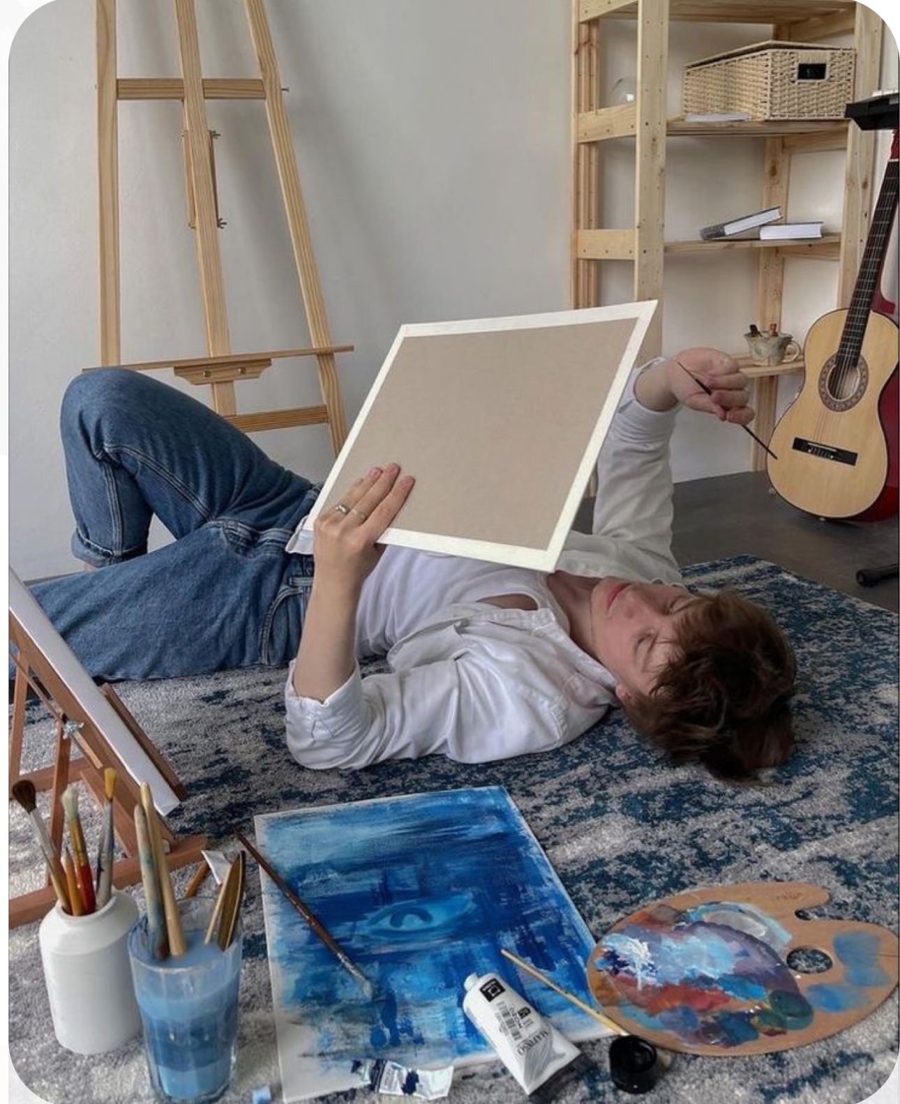 a young man lies on a blue rug in an art studio. he is holding a blank canvas in front of his face, obscuring his view.  surrounding him are art supplies including a wooden easel with a partially finished blue painting on it, paintbrushes in a jar, a palette of paint, and tubes of paint. there is a wooden shelf unit behind him holding books, art supplies, and a guitar. the overall feel of the image is artistic and serene.