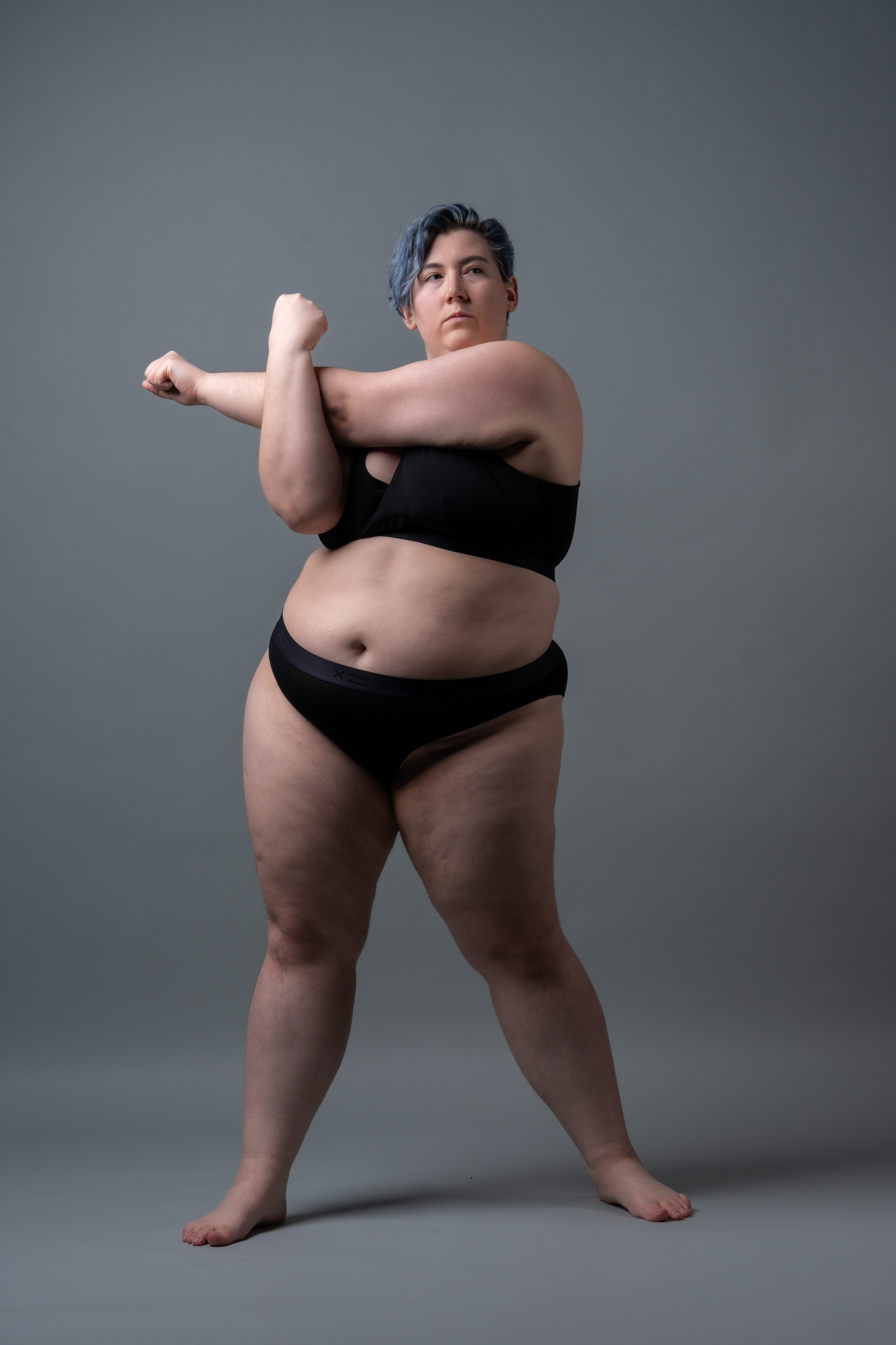 a plus size woman with short blue hair wearing black underwear is stretching with her arms crossed in front of a gray background.
