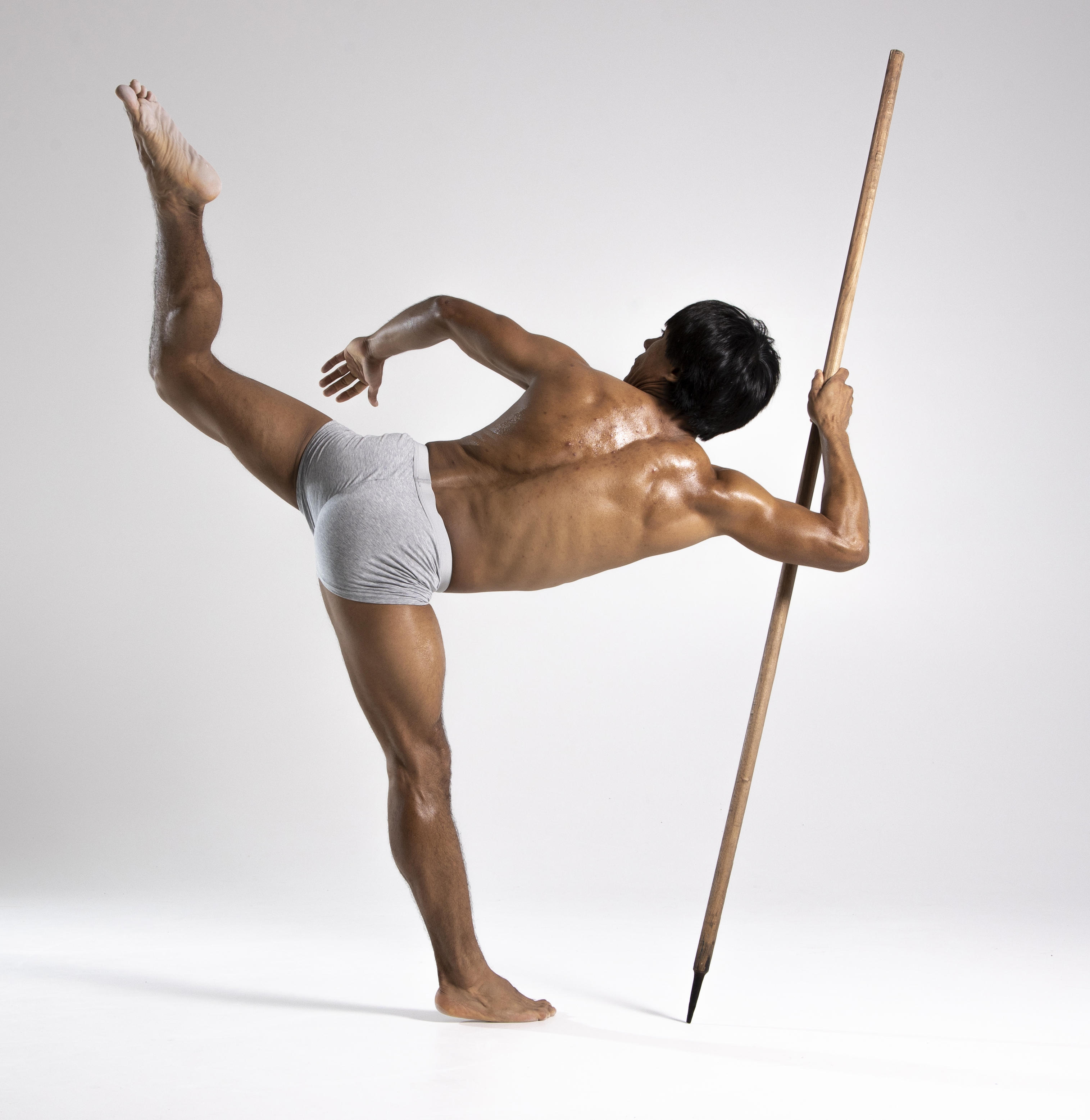 a muscular man, possibly a dancer or bodybuilder, is striking a pose in a studio. he is barefoot and wearing gray shorts. he has one leg extended high in the air, while the other leg is supporting his weight. he is holding a wooden pole for balance. his skin appears to be oiled. the background is a plain white. he is seen from the rear.
