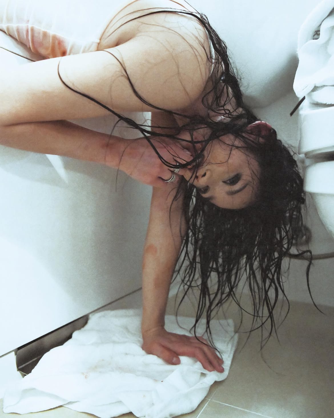 a woman with wet, dark hair is bent over in a bathroom, upside down, with her buttocks exposed. she is using a white towel to support her weight, and there are bruises visible on her arm. the background shows a toilet and tiled walls.