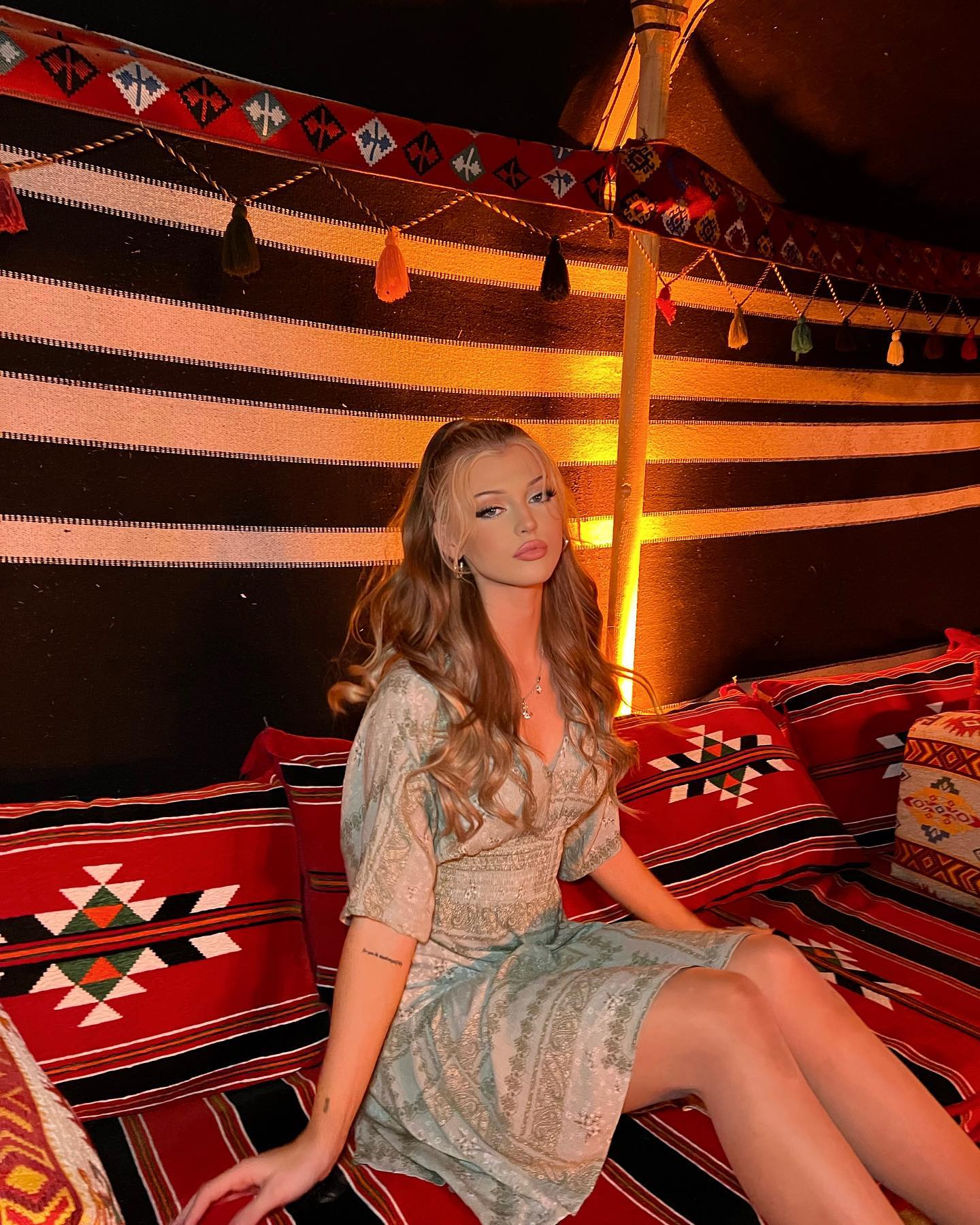 a woman with blonde hair wearing a dress is sitting in a tent. the tent is decorated with patterned fabric and tassel decorations. pillows with geometric patterns are arranged on the seating area.