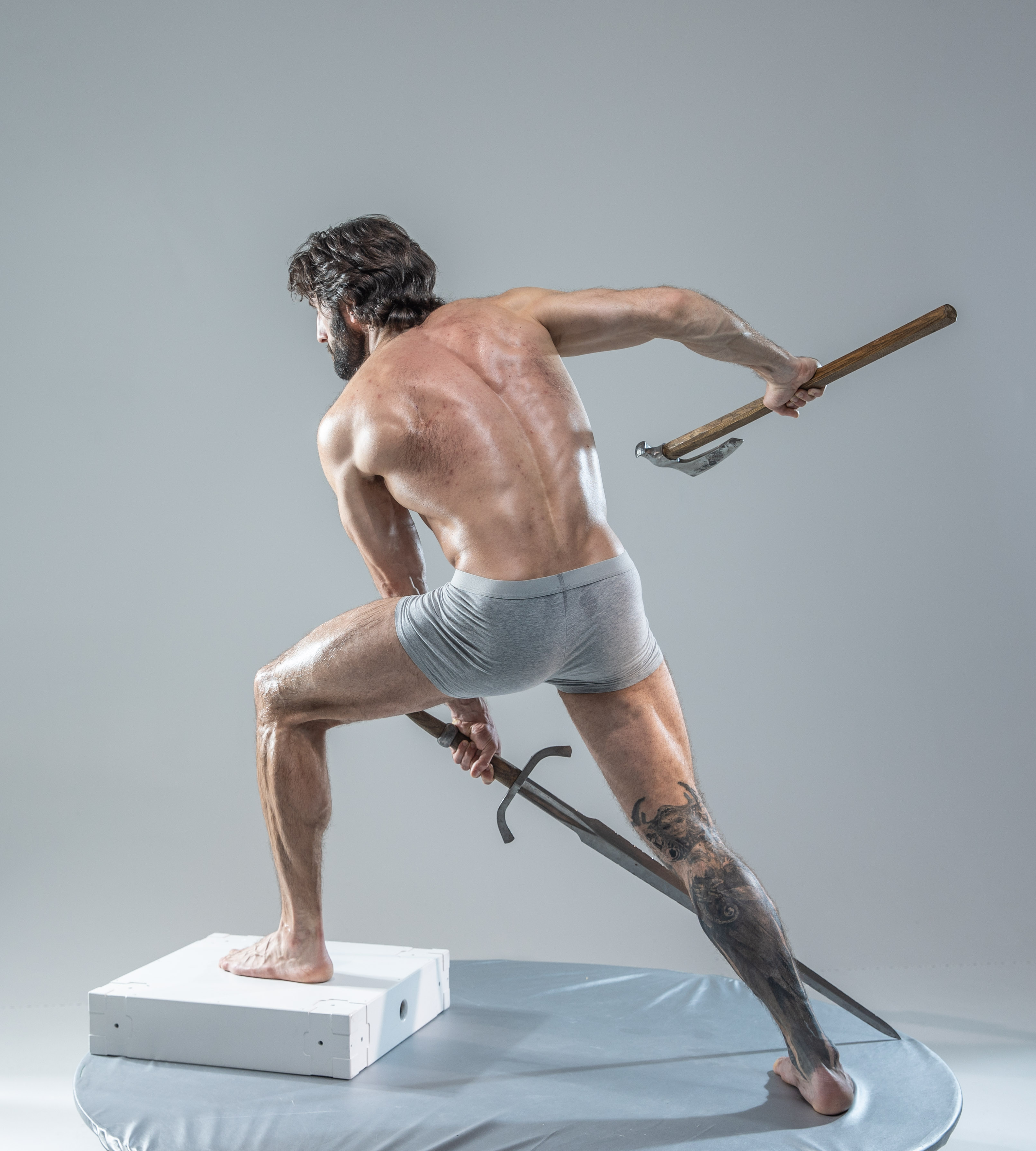 a muscular, shirtless man with a beard stands on a white platform in a fighting stance. he is holding an axe in his right hand, raised above his head, and a sword in his left hand, pointed downward. he is wearing gray underwear and has a tattoo on his right leg. the background is a light gray, and the lighting is soft.  the overall image emphasizes the man's physique and the power of his pose.