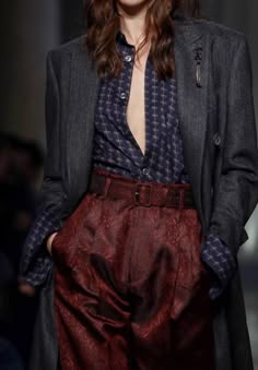 a woman is posing in a fashionable outfit.  she is wearing a red and black patterned shirt with a grey jacket over it. she has burgundy colored pants on. the woman's hair is long and styled and she is wearing silver accessories. the woman is posing with a confident stance and a relaxed expression.  she appears to be in a photoshoot.