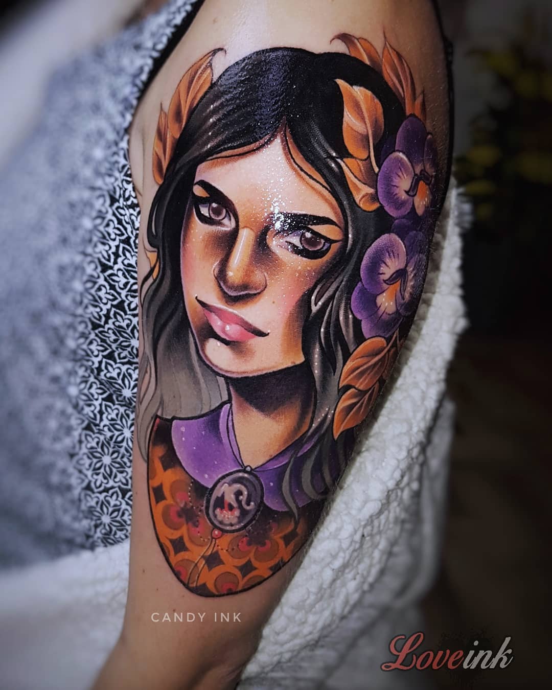 a neotraditional tattoo on someone’s upper arm. the tattoo is a portrait of a woman with dark hair, wearing a gold and purple patterned top, a white shirt underneath, and a necklace with a skull pendant. there are purple flowers and orange and brown leaves behind her. the background of the image is out of focus and appears to show a person wearing a fuzzy white sweater. the words “candy ink” are written in white, capitalized letters, and small font over the person’s arm. in the bottom right corner of the image, in cursive and stylized pink letters with a pink drop shadow, are the words “love ink”.