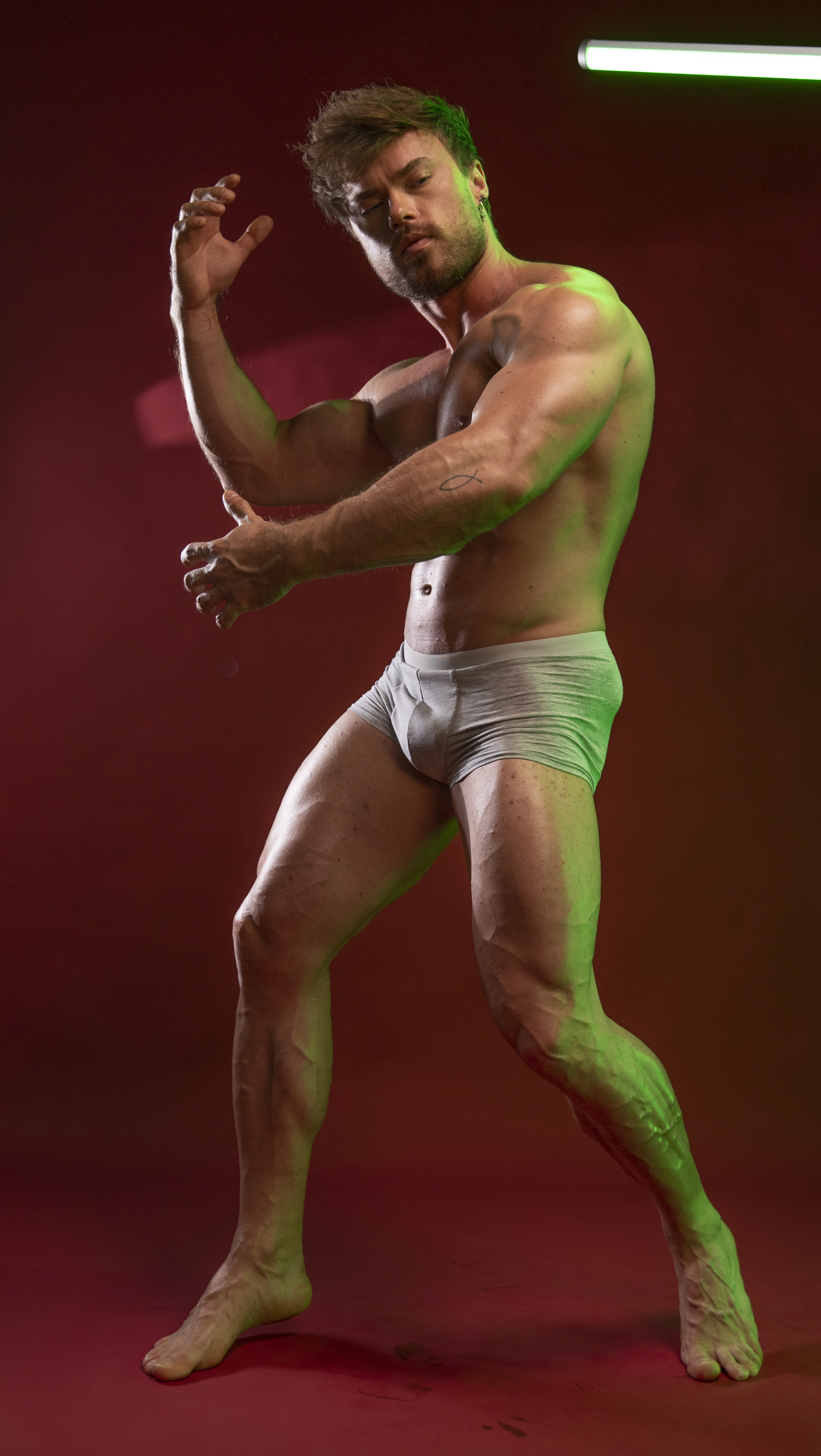 a shirtless, muscular man wearing underwear is posing in a studio setting. he has a bodybuilder physique with defined muscles and visible veins, especially in his arms and legs. the background is red, and there is green lighting illuminating his body, creating a dramatic effect. he is barefoot and positioned in a dynamic stance.