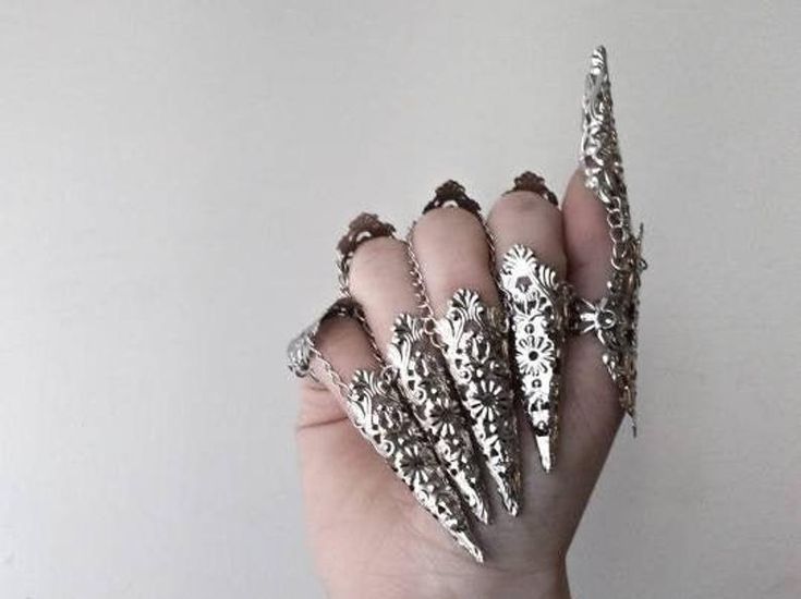 a hand is adorned with ornate, silvercolored metal nail jewelry on each finger. the jewelry resembles pointed claws with intricate filigree designs. the hand is in front of a light gray background.