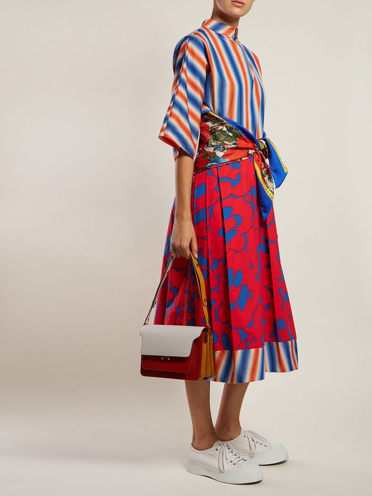 a fullbody shot of a woman modeling a stylish outfit. she's wearing a red pleated midi skirt with a bold floral print, paired with a shortsleeved striped shirt in shades of blue, orange, and white. a vibrant blue and green scarf is tied around her waist, adding a pop of color. the outfit is completed with a multicolored (grey, red, and yellow) crossbody bag and white sneakers.  the model is standing against a plain white background.