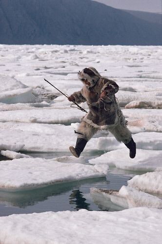 a man dressed in traditional inuit fur clothing jumps across ice floes in an arctic environment, holding a spear, likely hunting. the scene is dominated by the icy landscape, with snowcovered ice floes surrounding open water, and a distant mountain range in the background.