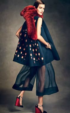 a fashionable young woman in a blue dress, black sheer pants, red fur collar, and high heels poses in a studio setting. she is facing the camera with a slight smile.