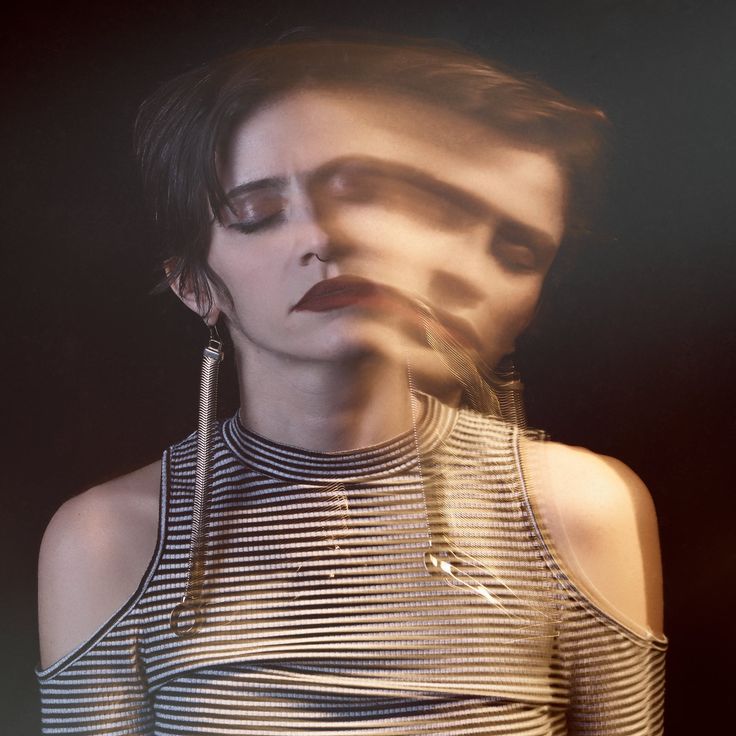 a woman with dark hair wears a black and white striped, cold shoulder top, and silver earrings. her eyes are closed and she is staring downwards with a thoughtful expression. the image is shot in a dark studio and has a moody, dramatic aesthetic.