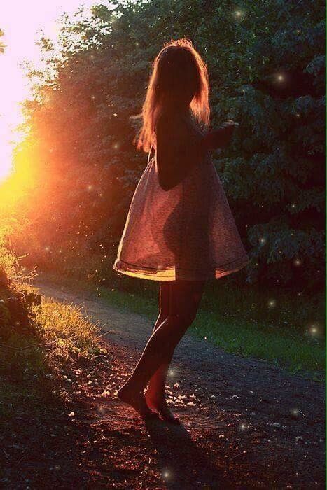 a silhouette of a young woman walking barefoot on a path in a forest at sunset. she is wearing a long, flowing dress and her hair is long and flowing. the light from the setting sun creates a magical, dreamy atmosphere. there are sparkles of light in the air.