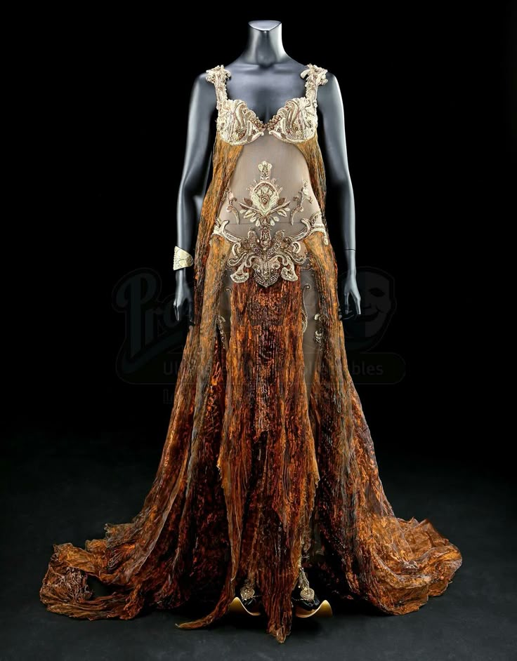 an ornate, bronze and gold gown is displayed on a mannequin. the top of the dress features an elaborately decorated bustierstyle top and a sheer fabric panel with embroidered designs. the lower portion of the gown has a textured, flowing fabric that mimics wood grain. it is presented on a black background with some branding imagery in the background.