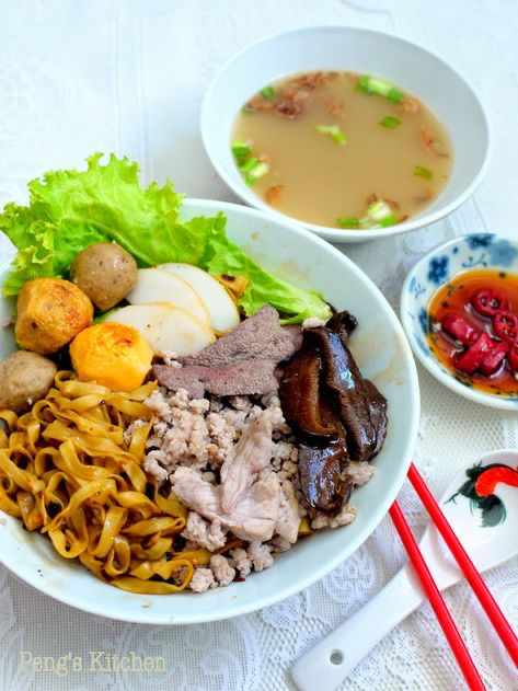 a closeup shot of a bowl of chinese noodles with various toppings, including meat, dumplings, and an egg. a bowl of soup, chopsticks, and a small dish of chili sauce are also visible in the image. the food is presented in a visually appealing manner, making it look delicious. the background is a white tablecloth, which contrasts with the vibrant colors of the food.