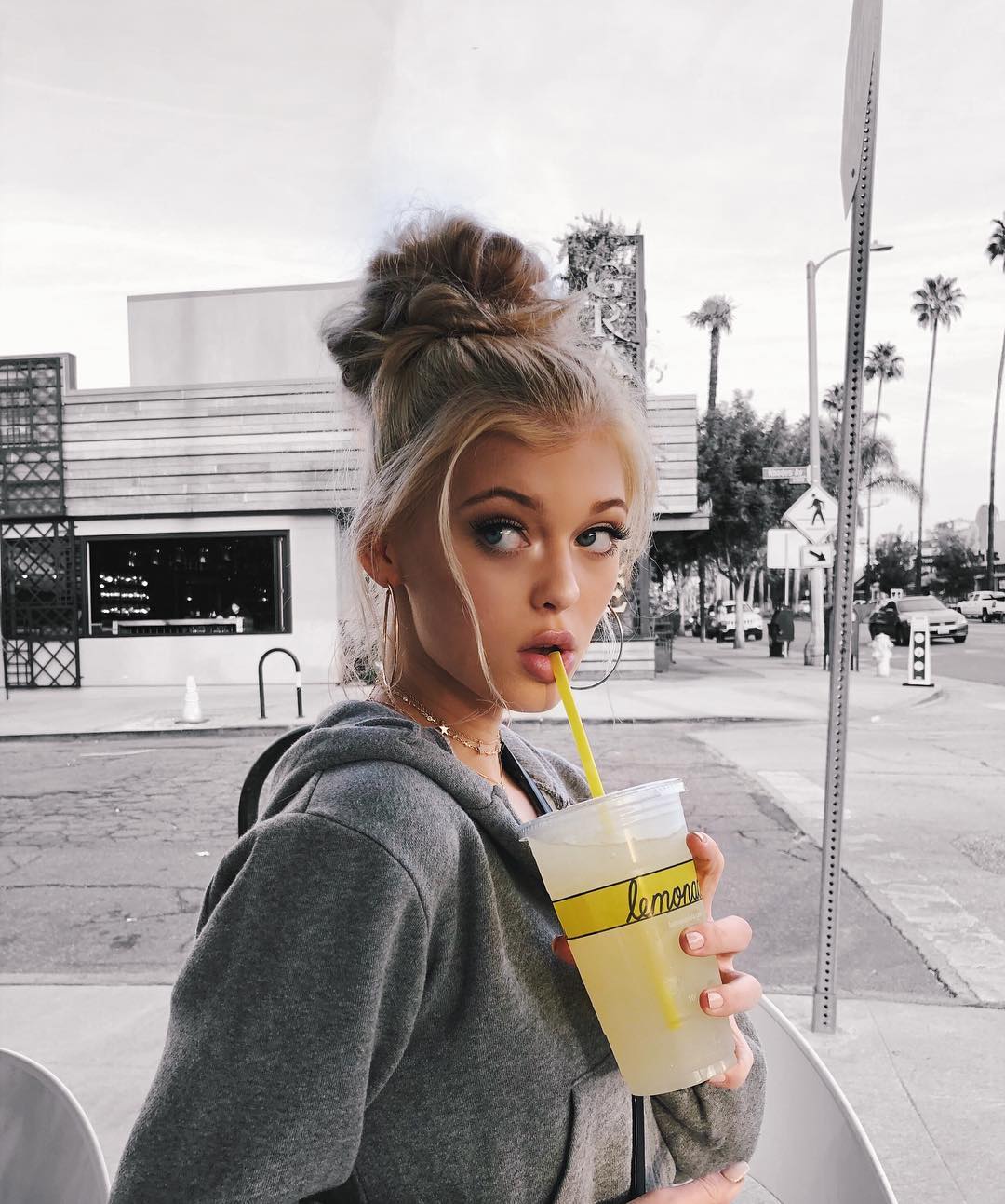 a woman with blonde hair in a bun wearing a gray hoodie and gold hoops is drinking lemonade with a straw. she is on the street near a building and palm trees. the image is in gray scale.
