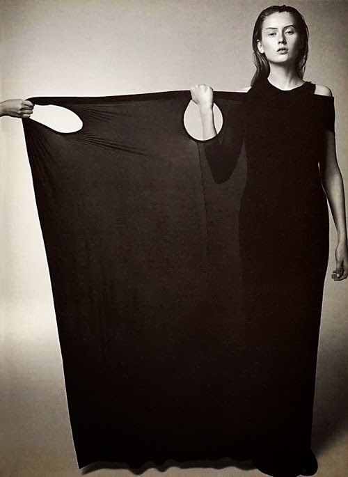 a woman in a black dress stands holding a large, dark fabric with two circular holes in it. the fabric is stretched out, revealing her hands holding the edges. the dress has a shoulder cutout. the image appears to be a studio shot with a neutral background.