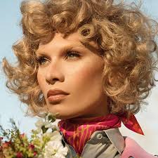 closeup portrait of a beautiful young woman with short, curly blonde hair. she's wearing a pink and orange floral print silk scarf tied around her neck, a light beige collared shirt, and possibly a jacket. she is outdoors, bathed in sunlight, with a bouquet of flowers partially visible in the foreground. her makeup is natural and enhances her features, creating a fresh and healthy look.  the overall impression is one of elegance and serenity.