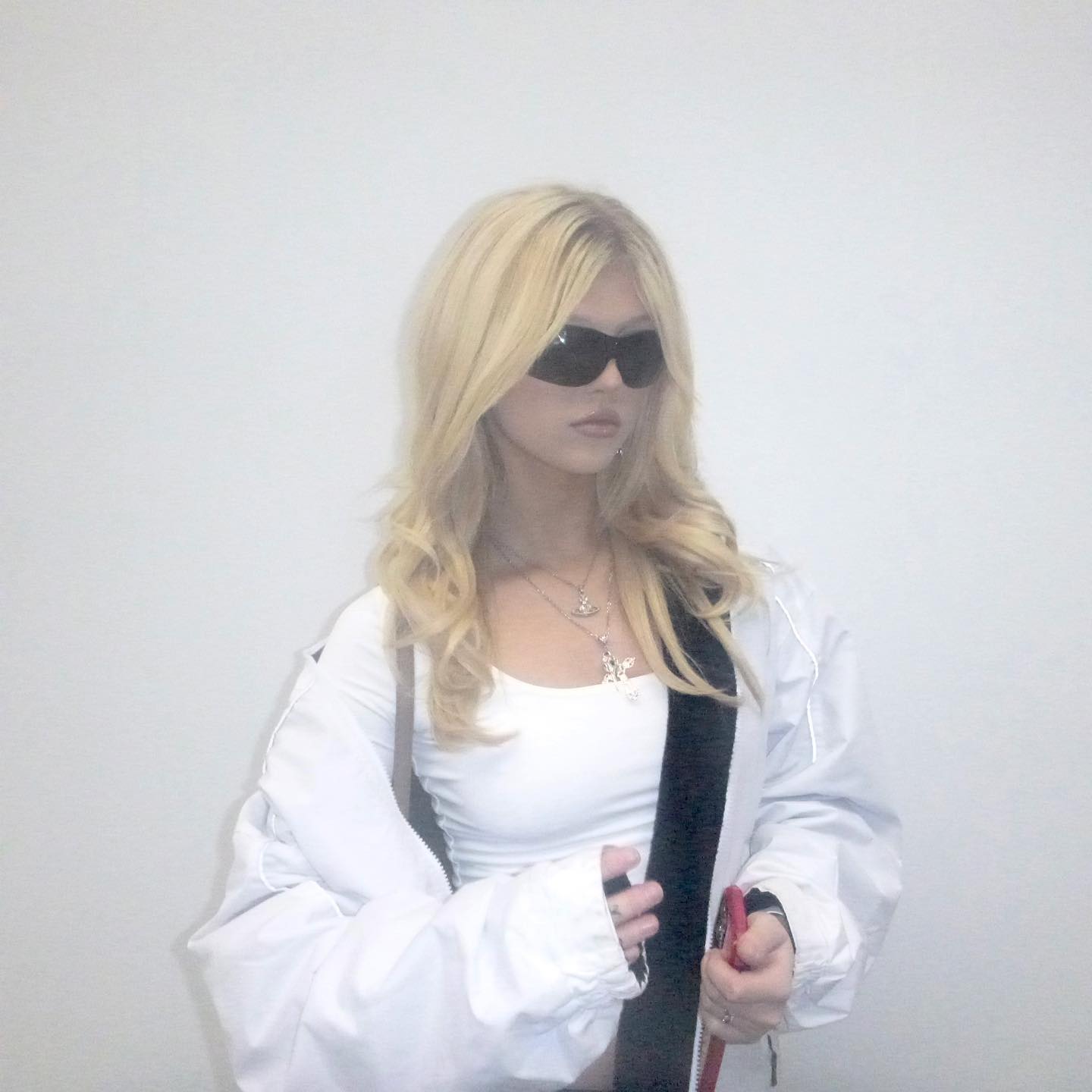 a woman with blonde hair wearing sunglasses and a white top under a white jacket is posing indoors. she is also wearing jewelry, including a necklace.
