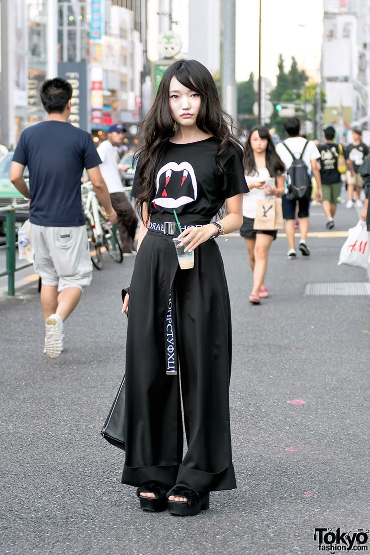 a stylish asian woman is standing on a city street, dressed in a black tshirt with a graphic of lips with fangs, wideleg black pants with a belt detail, and black platform shoes. she is holding a straw cup. the background shows a busy urban setting with pedestrians and buildings, consistent with tokyo fashion street style.
