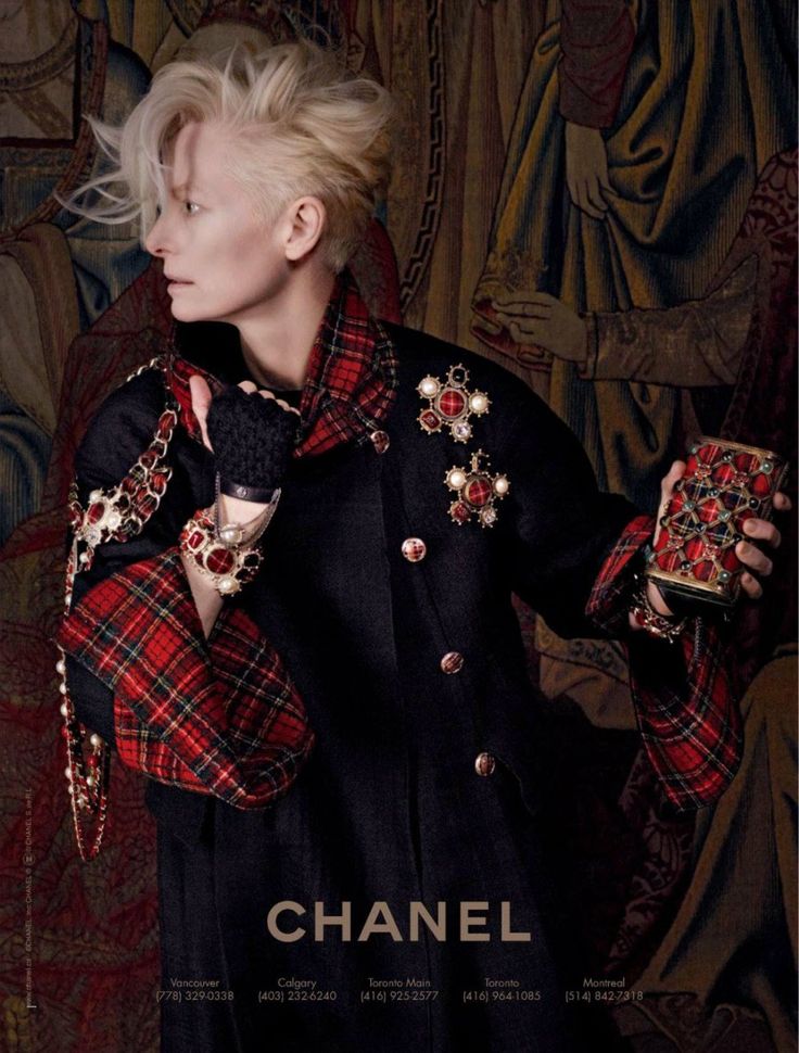a woman with short blonde hair is wearing a black coat with a red and black plaid scarf, chain jewelry, and gloves. she is holding a small clutch bag and posing in front of a tapestry. the image is an advertisement for chanel.