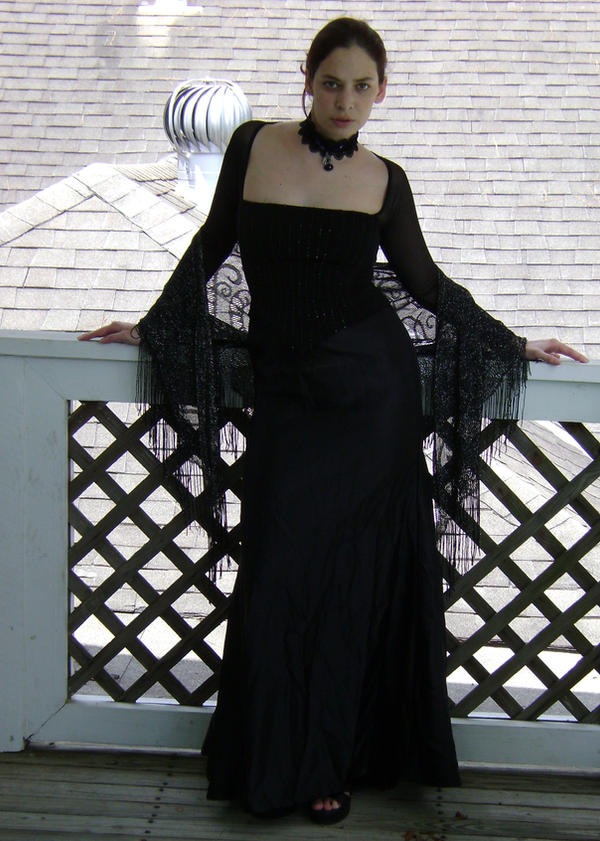 a woman is standing on a balcony in a long, black dress with a square neckline, long sleeves with tassel details, and a black choker. she is leaning against the white railing of the balcony, with a tiled roof and a weather vane in the background.
