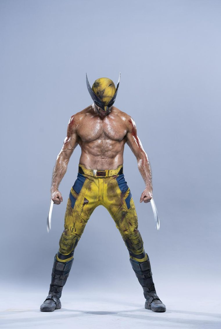 a full body shot of wolverine, the marvel superhero, standing with his claws extended. he's wearing his classic yellow and blue costume and mask, and his muscular physique is visible. he appears battleworn with some blood and dirt on his body and costume. the background is a light gray.